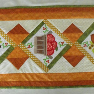 May include: A white table runner with a diamond pattern in orange, green, and yellow. The center of the runner features three embroidered images: a pumpkin, a basket of apples, and acorns.