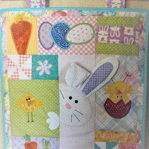May include: A colorful patchwork quilt with an Easter theme. It features a bunny rabbit, a chick, carrots, and eggs. The quilt is made of various fabrics in shades of pink, blue, green, yellow, and white.