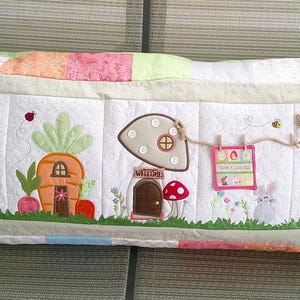 May include: A long, rectangular pillow with a white background and colorful patchwork fabric on the ends. The pillow features embroidered images of a carrot house, a mushroom house, a bunny in a boot, and a sign that says "Hoppin' Down the Bunny Trail".