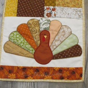 May include: A Thanksgiving table runner with a brown and white patchwork turkey design. The turkey has a brown body and a white head with a red wattle. The runner has a brown, white, and gold patchwork design with a brown and white polka dot pattern. The runner has a yellow border with a gold trim. The runner has the word "thankful" embroidered in brown thread on the bottom.