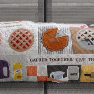 May include: A white quilted pillow with a Thanksgiving theme. The pillow features various embroidered images of pies, kitchen tools, and the text "Gather Together, Give Thanks, Eat Pie".