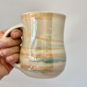 May include: A ceramic mug with a glossy, iridescent finish featuring horizontal bands of soft pastel colors including peach, light blue, and cream. The mug has a rounded body and a curved handle, with a smooth, reflective surface texture.