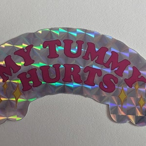Puede incluir: Una pegatina holográfica plateada con el texto "My Tummy Hurts" en letras rosas. La pegatina tiene dos estrellas doradas brillantes a cada lado del texto.