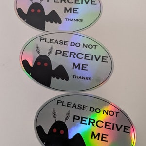 Puede incluir: Tres pegatinas hologr&aacute;ficas de forma ovalada con una ilustraci&oacute;n en blanco y negro de una criatura con ojos rojos y el texto "PLEASE DO NOT PERCEIVE ME THANKS".