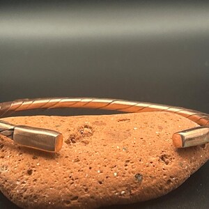 May include: A copper cuff bracelet featuring a twisted, braided metal design along the band and smooth, rectangular end caps. The metallic surface has a warm, reddish-brown tone with visible highlights reflecting light along the twisted texture.