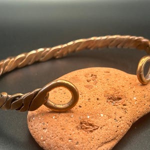 May include: A copper-colored metal cuff bracelet featuring a twisted, braided design throughout the band. The bracelet ends in two circular loops that form the opening of the piece, showcasing a metallic finish with subtle variations in tone.