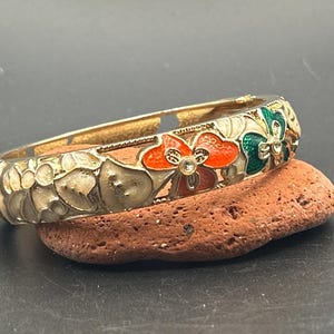 May include: A gold-tone hinged bangle bracelet featuring an enamel floral pattern in shades of cream, orange, and green. The design includes small clear rhinestones set in the center of the flowers, with a polished metal interior finish.