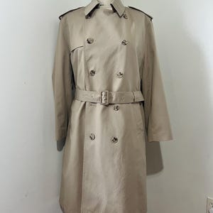 May include: A tan trench coat with a double-breasted design, featuring a belt at the waist and button closures. The coat is displayed on a mannequin, showcasing its classic style and tailored fit. The coat has a collar and shoulder epaulets.