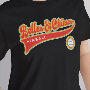 Puede incluir: Camiseta negra con el texto "Belles & Chimes PINBALL" en un diseño retro. El diseño presenta letras rojas y amarillas y un pequeño logotipo circular. La camiseta es de cuello redondo y manga corta.