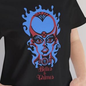 以下が含まれることがあります： 赤と青の様式化された顔のデザインと「Belles & Chimes」の文字が入った黒いTシャツ。 グラフィックは炎のようなアウトラインです。 Tシャツはクルーネックです。