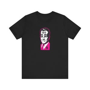 May include: Black t-shirt featuring a pop art style portrait of a man in a pink suit with a barcode over his head. The design includes a dollar sign on the suit. The graphic is in pink and white.