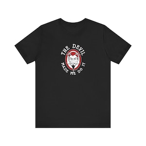 Peut inclure: T-shirt noir avec un cercle rouge et du texte blanc qui dit "The Devil Made Me Do It". À l'intérieur du cercle se trouve une image de dessin animé d'un diable avec des cornes et une barbiche.