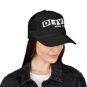 Könnte beinhalten: Eine schwarze Trucker-Cap mit Netzrückseite und festem Vorderteil. Auf der Vorderseite befindet sich der weiße Schriftzug DEVO in einer fetten, blockartigen Schrift, darunter die Worte was right in einer kleineren, serifenlosen Schrift.