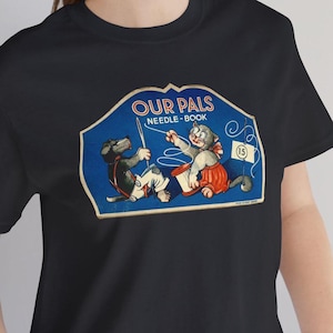 May include: Black t-shirt with a vintage illustration of a cat and dog holding a spool of thread. The illustration is surrounded by a blue border and the text "Our Pals Needle-Book 15" is printed on the border.