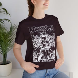 May include: Black t-shirt featuring a white graphic of a comic book-style illustration with the text "Alien Star" and "A Gottlieb Pinball Game". The graphic depicts several figures in armor.