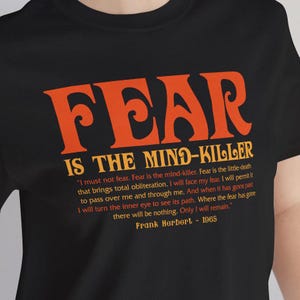 May include: Black t-shirt with the orange text "FEAR IS THE MIND-KILLER". The quote by Frank Herbert from 1965 is also printed on the shirt. The shirt is a crew neck.