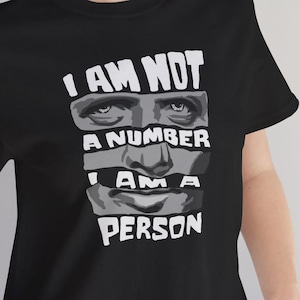 May include: Black t-shirt with a graphic design featuring a fragmented face and the text "I AM NOT A NUMBER I AM A PERSON" in white. The design is a bold statement.
