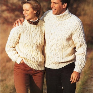 May include: A couple wearing white cable knit sweaters with a turtleneck and a crew neck. The woman is wearing brown trousers and the man is wearing black trousers.