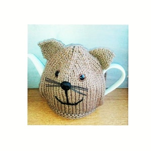 Op de afbeelding: Een bruine gebreide theepotwarmer in de vorm van een kattengezicht met zwarte ogen, een zwarte neus en zwarte snorharen.