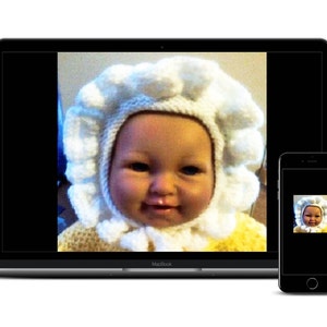 May include: A baby wearing a white crocheted hat with a ruffled edge. The hat has a white crocheted hood that covers the baby's ears and neck. The baby is smiling and looking at the camera.