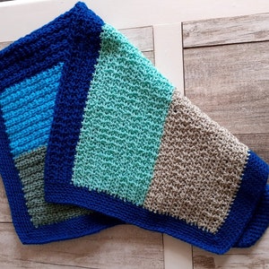 May include: A crocheted baby blanket in a blue, teal, grey and white colour scheme. The blanket is folded in half and has a blue border.