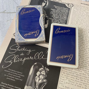 May include: Two decks of "Bonsoir" playing cards with navy blue backs and gold lettering, presented on a vintage magazine page. The cards are in their original boxes, with a blue and white floral design.