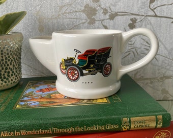 May include: White ceramic shaving mug with a black, red, and yellow vintage car illustration and the word "FORD". The mug has a handle and a spout. It is sitting on top of a stack of books.
