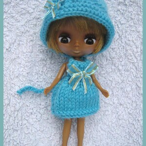 May include: A doll wearing a handmade blue knitted dress and matching hat with a bow. The doll has brown skin, brown eyes, and short blonde hair. The hat has a small bow with gold accents.