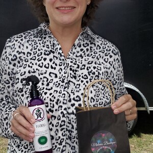 Op de afbeelding: Een vrouw met een wit en zwart luipaardprint shirt houdt een paarse fles haarproduct vast met het label "FAB'U" en een zwarte boodschappentas met een sticker waarop staat "Malena Boutique - Een beetje van alles".
