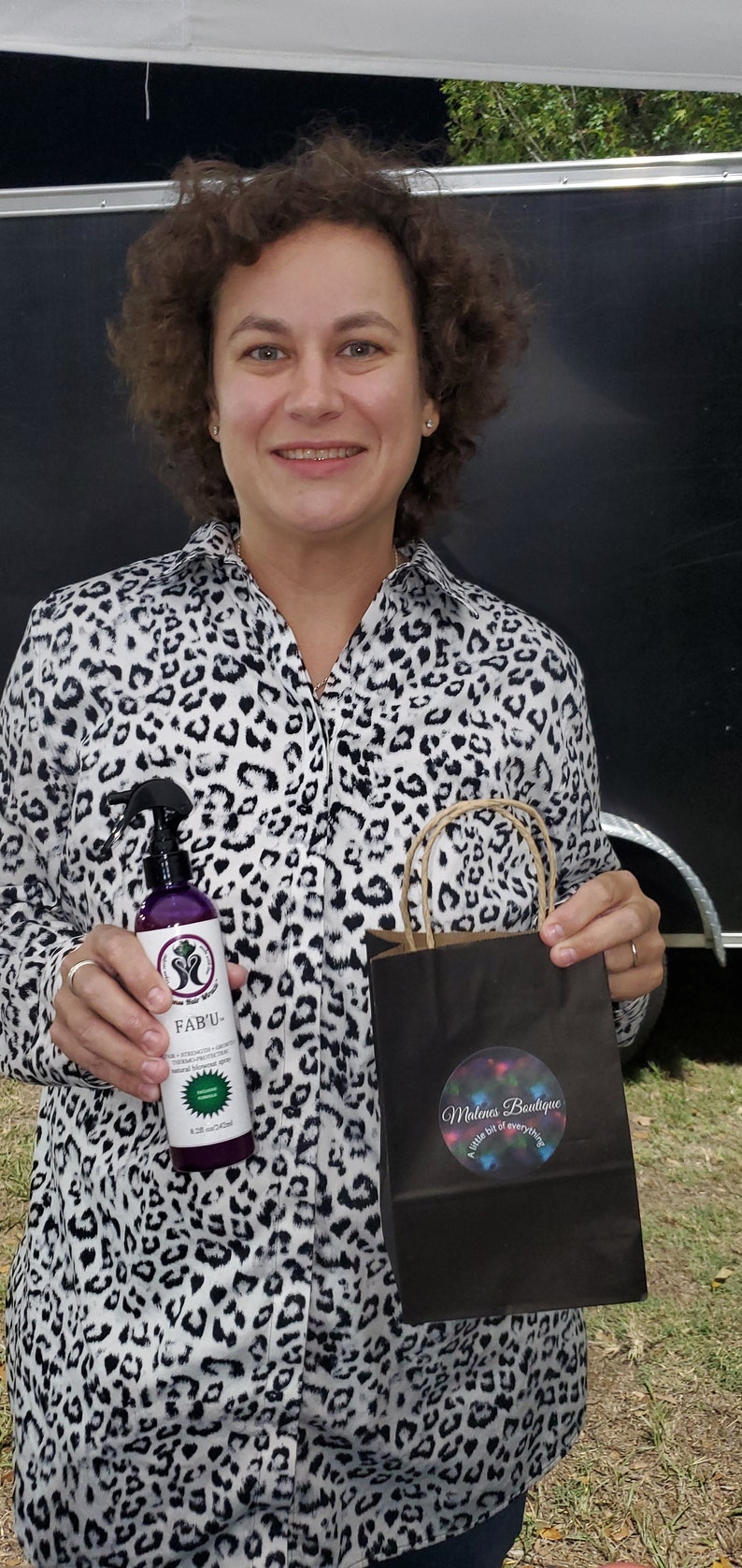 Op de afbeelding: Een vrouw met een wit en zwart luipaardprint shirt houdt een paarse fles haarproduct vast met het label "FAB'U" en een zwarte boodschappentas met een sticker waarop staat "Malena Boutique - Een beetje van alles".