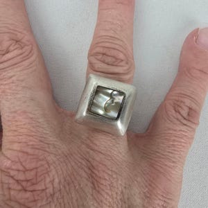 Peut inclure: Une bague de forme carrée avec une finition argentée mate, dotée d'une incrustation carrée centrale en nacre irisée. La bague présente une bande épaisse et substantielle ainsi qu'un design aux bords arrondis entourant le centre en coquillage.