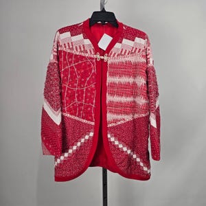 Peut inclure: Une veste à manches longues présentant un motif patchwork dans des tons de rouge et de blanc. Le vêtement possède une encolure en V, une fermeture à bascule de couleur claire sur la poitrine et une texture matelassée avec divers motifs.