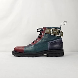 May include: A multi-coloured leather ankle boot featuring a red toe cap, teal side panels, a dark purple heel section, and a maroon lace-up front. The boot includes a green ankle strap with a silver metal buckle and a thick black lugged sole.
