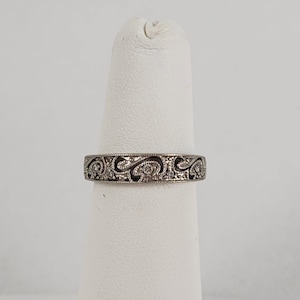 May include: A silver-toned metal ring featuring an intricate, repeating scrollwork pattern across the band. The design includes small, embedded clear stones set within the recessed, darkened areas of the metal, creating a textured, detailed appearance.