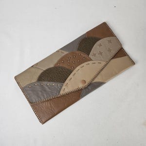 May include: A rectangular clutch wallet featuring a patchwork design of various leather textures in shades of brown, tan, and grey. The exterior is accented with decorative whipstitching along the seams and a single snap button closure.
