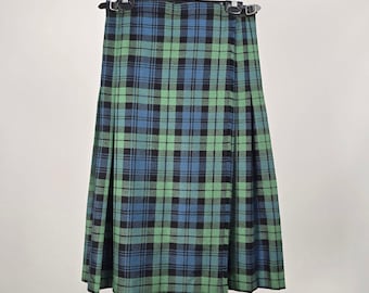 Peut inclure: Un kilt plissé avec un motif à carreaux dans des tons de vert, bleu et noir. Le vêtement comprend deux sangles réglables en cuir noir avec des boucles en métal argenté à la taille, suspendu à un cintre en plastique noir sur fond uni.
