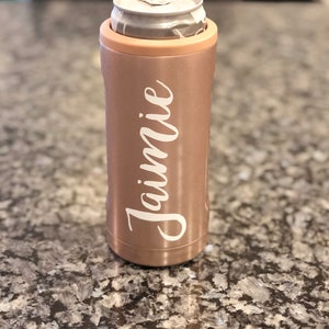 May include: A rose gold can cooler with the name "Jamie" written in white cursive on the side. The cooler is holding a silver can.