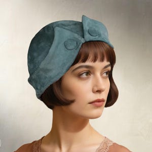 Può includere: Un cappello cloche in camoscio color teal con due bottoni decorativi. Il cappello ha una tesa curva e una corona arrotondata. Il testo "Elschen Sewing Pattern" e "Instant PDF Download" sono visibili.
