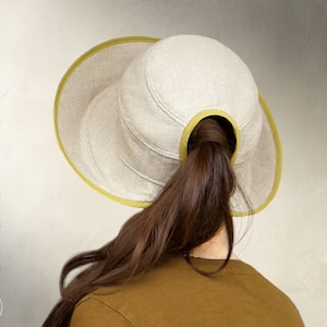 May include: A light beige linen hat with a yellow trim and a hole in the back for a ponytail. The hat is worn by a person with long brown hair.