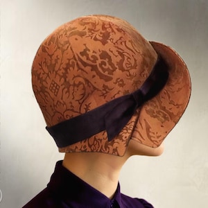 May include: A vintage-style cloche hat in shades of brown with a dark purple ribbon. The hat features a detailed, all-over pattern. The words "Elsewhen Sewing Pattern" and "Instant PDF Download" are printed vertically on the sides.