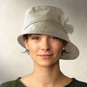 Op de afbeelding: Een lichtgrijze cloche hoed met een licht asymmetrische rand en een delicate stoffen bloem. De hoed is gemaakt van een textuurstof en wordt door een persoon gedragen. De woorden "Elsenhen Sewing Pattern" en "Instant PDF Download" staan op de zijkanten.