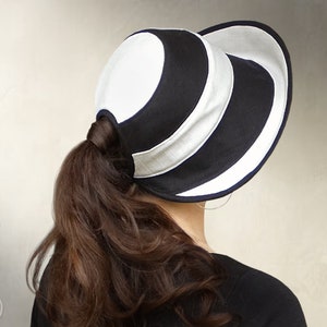 Può includere: Un cappello da sole a righe bianche e nere con un'ampia tesa. Il cappello ha un foro sul retro per una coda di cavallo.