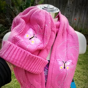 May include: Pink knitted hat and scarf set with embroidered butterflies. The hat and scarf are both decorated with rhinestones.