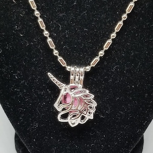 May include: A silver unicorn charm necklace with a pink and white unicorn inside the charm. The necklace is on a black velvet stand. There is a small pink lava rock next to the necklace.