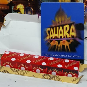 May include: A close-up of six red dice with gold lettering that reads "Sahara" and "Las Vegas". The dice are arranged in a row and are sitting on a gold and brown surface. A blue card with the words "Sahara Hotel and Casino, Las Vegas" is visible in the background.