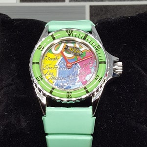 May include: A green wristwatch with a silver band and a map of the United States on the face. The watch face also has the text "USPS" on it.