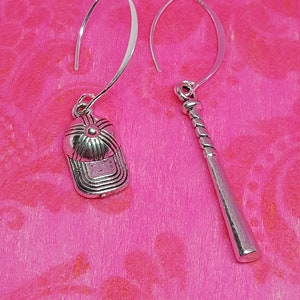 May include: A pair of silver earrings with a baseball cap and a baseball bat charm. The earrings are hanging from a pink background.