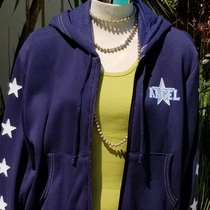 May include: A navy blue zip-up hoodie with white star accents on the sleeves and the word "ANGEL" in a starburst design. The hoodie is paired with a light green tank top and a denim mini skirt with metal grommet details.