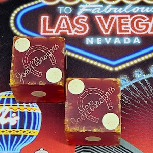 May include: Two red dice with white dots and the text "Joel Brown's" in gold lettering. The dice are in front of a neon sign that says "Welcome to Fabulous Las Vegas, Nevada".
