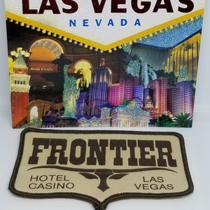 May include: A tan-colored patch with brown stitching and the text "FRONTIER HOTEL CASINO LAS VEGAS" in brown lettering. The patch features a stylized brown bull's head.